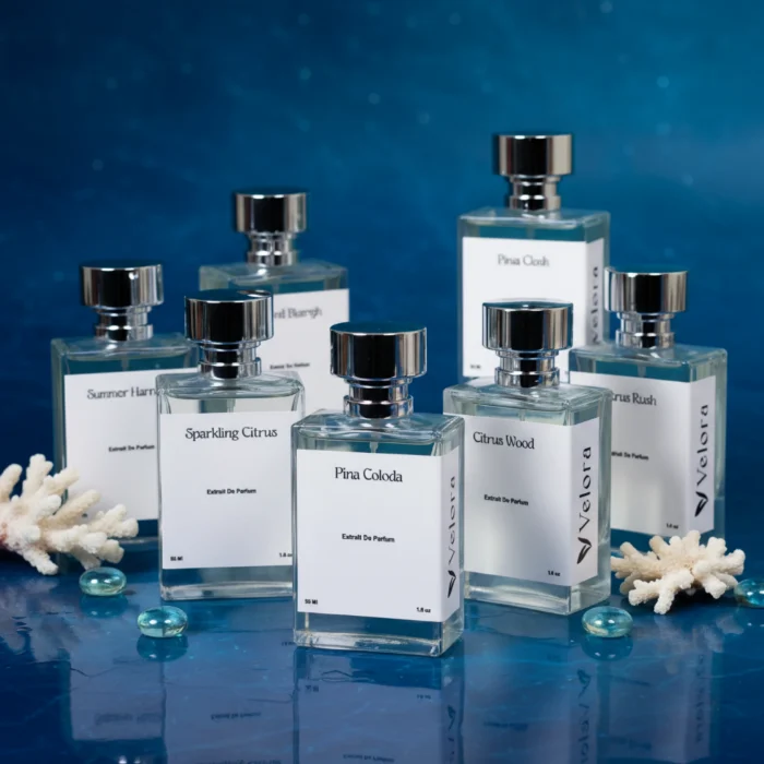 مجموعة عطور Velora على سطح أزرق عاكس مع مرجان أبيض وحصى زجاجي، ومنها Sparkling Citrus وPina Colada وCitrus Wood